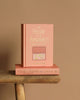 Two "Hound of the Baskervilles | Collector's Edition | Hardcover" books by Sir Arthur Conan Doyle are shown on a brown background. One stands upright, and the other lies flat on wood; both have pink covers with gold lettering, capturing Sherlock Holmes' mystery allure.