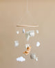 A nursery Handmade Mobile - Honey Bear - Final Sale hanging from the ceiling, featuring a plush teddy bear, white clouds, fluffy sheep, and embroidered cushions with words like "funny" and "happy" on a neutral background.