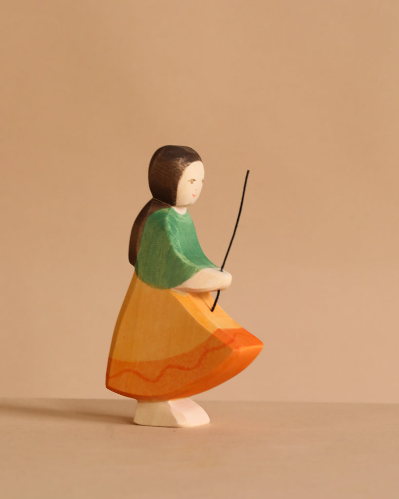 The Ostheimer Goose Girl is a handcrafted wooden figurine featuring a girl with long dark hair in a green top and orange-yellow skirt, holding a thin black stick—ideal for fans of non-toxic Ostheimer toys.