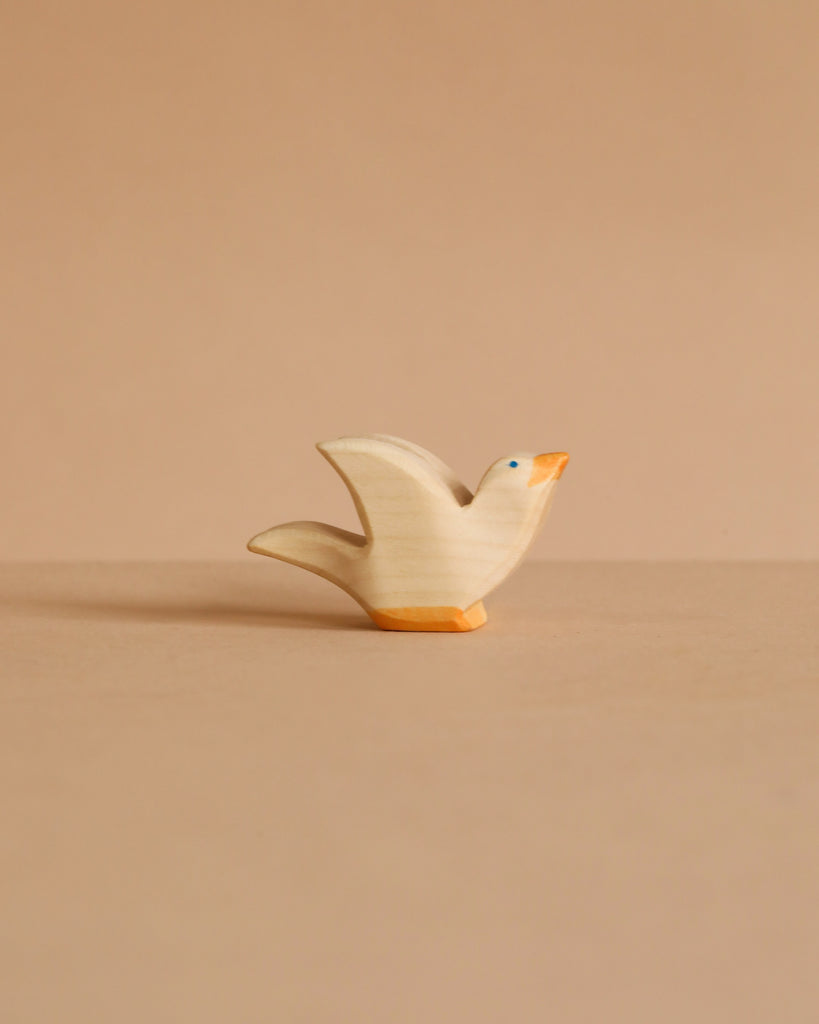 The Ostheimer Flying Pigeon is a small wooden figurine with an orange beak and base, handcrafted from non-toxic wood, shown against a plain beige background.