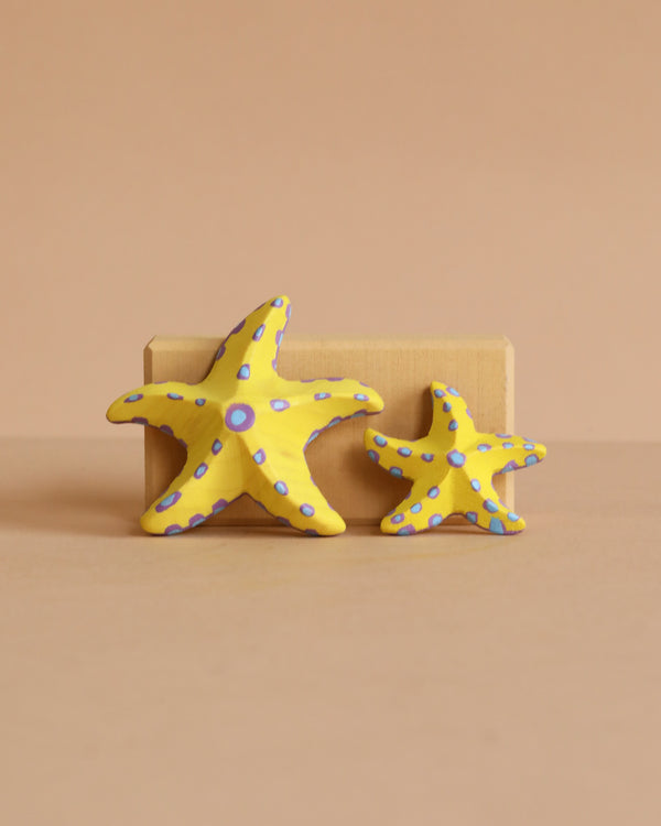 The Bumbu Handmade Wooden Yellow Starfish Set features two upright yellow wooden starfish with blue dots—one large, both with five arms—displayed against a beige block, making them ideal Montessori toys for creative, hands-on learning.