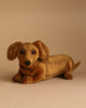 A realistic Dachshund Puppy Dog Stuffed Animal lies on a beige surface. The toy dog has caramel-brown fur, floppy ears, small black eyes, and a black nose. Hand-sewn by HANSA animals, the neutral background highlights the lifelike features of this plush toy.