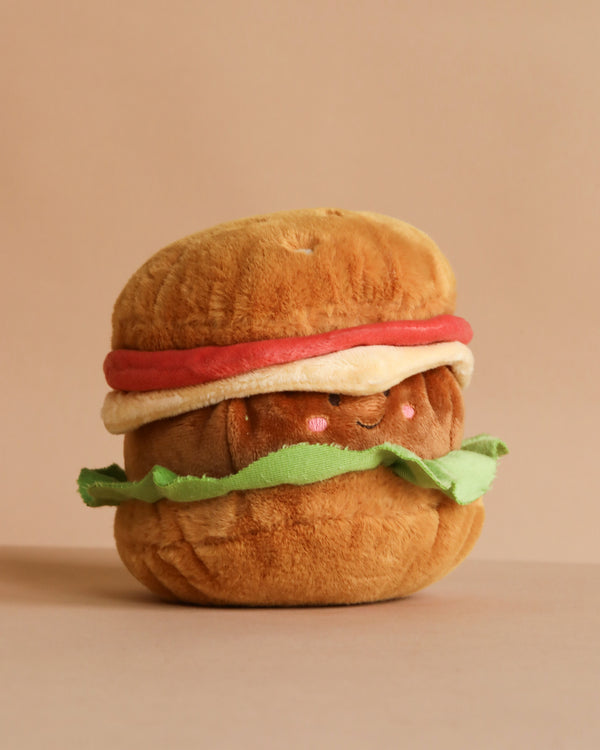The MON AMI Joe Burger Plush Toy features a smiling hamburger design with pink cheeks, tomato, lettuce, and a bun—making it perfect as cute decor for any space. Set against a plain beige background.