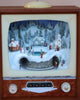 The Christmas Music TV With Train In Tunnel showcases a vintage TV frame displaying a winter village scene with snow-covered houses, pine trees, a train track, and tiny figurines. Sparkling snow adds a festive touch while Christmas songs play in the background, creating a charming holiday diorama. Please note that batteries are required for operation.