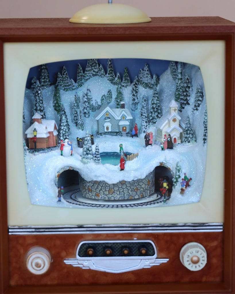 The Christmas Music TV With Train In Tunnel showcases a vintage TV frame displaying a winter village scene with snow-covered houses, pine trees, a train track, and tiny figurines. Sparkling snow adds a festive touch while Christmas songs play in the background, creating a charming holiday diorama. Please note that batteries are required for operation.