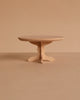 The PRE-ORDER: Maileg Oval Dining Table, Mouse Size - Nature features a small, round wooden design with a single pedestal base, displayed on a plain beige surface that highlights SuitCasa’s signature wood quality.