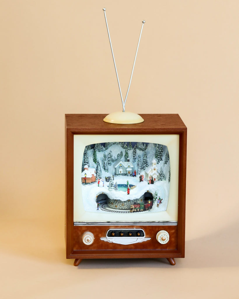 A Christmas Music TV With Train In Tunnel, styled like a vintage television complete with an antenna, evokes the warmth of holiday tunes as it showcases a miniature snowy village scene on the screen. The display highlights small houses, trees, and people, all set within a classic wooden design featuring dials. This charming piece stands against a neutral background.