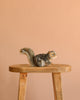 A Gray Squirrel Stuffed Animal, with a lifelike design reminiscent of hand-sewn toys, is displayed on a wooden stool against a plain peach-colored background.