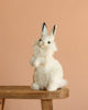 The White Bunny Stuffed Animal sits upright on a wooden stool against a peach background, highlighting its realistic plush design and quality craftsmanship.