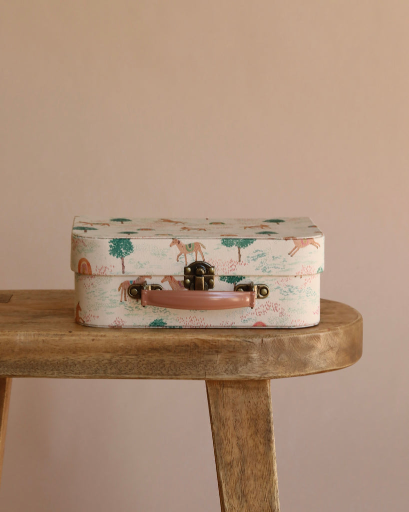 The Maileg On The Go Suitcase Set in mouse size, featuring a charming animal and nature print, rests closed on a wooden bench against a plain beige background.