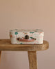 The Maileg On The Go Suitcase Set, featuring a whimsical animal and tree print in vintage style, is displayed on a wooden bench against a beige background—perfect for imaginative Maileg Mouse adventures.