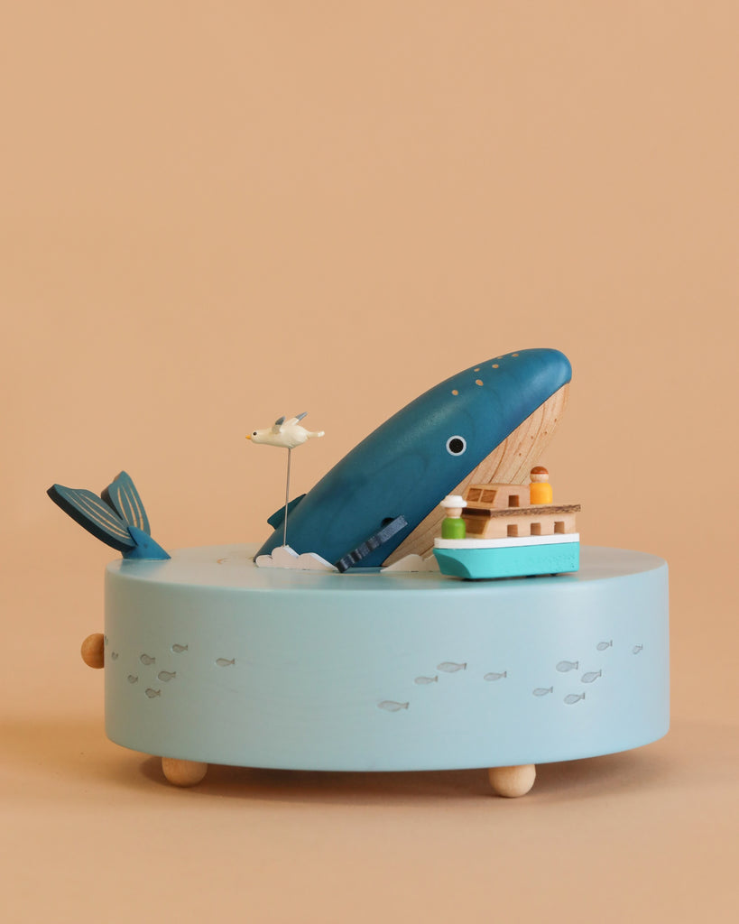 The Wooden Whale Music Box is a blue, ocean-themed wooden box with a whale, seagull, and small boat on top. Its round light blue base features tiny fish painted over a beige background.