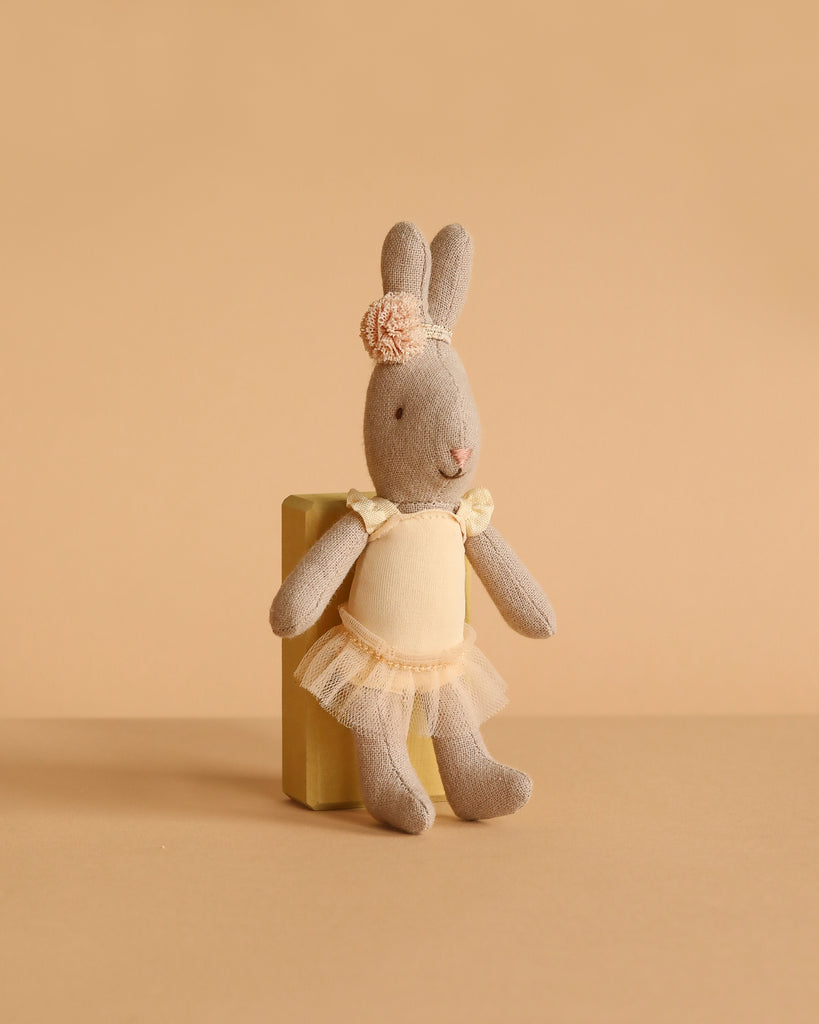 A Maileg Rabbit, Micro - Ballet Suit and Skirt, Cream (ships in one week), poses on a small yellow block against a beige background, resembling an adorable ballerina ready for the stage.