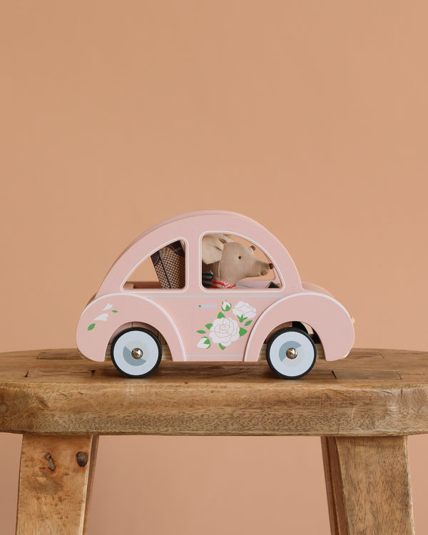 The Maileg Road Trip Adventure Set features a small toy mouse in a flower-adorned wooden car, perfect for dollhouses, all displayed on a rustic stool with a peach backdrop.