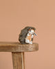 A 4-inch Steiff Joggi Baby Hedgehog Stuffed Plush Toy with a brown and white body and signature Button in Ear tag sits on the edge of a wooden stool, set against a plain peach-colored background.