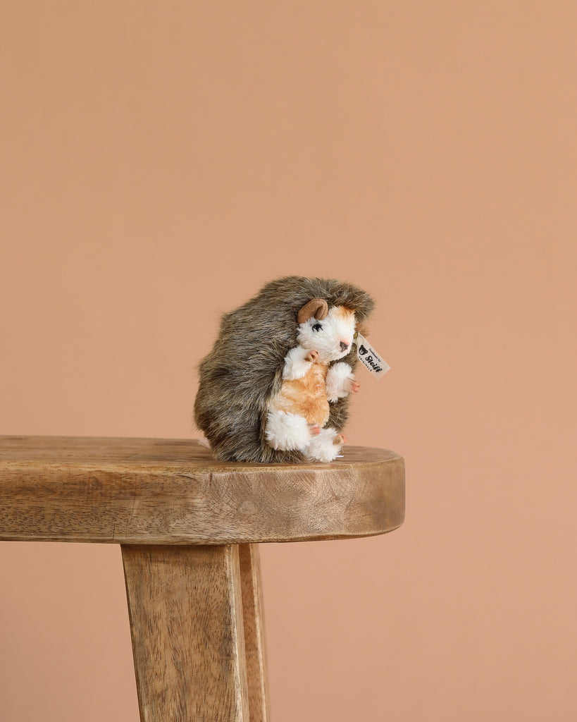 A 4-inch Steiff Joggi Baby Hedgehog Stuffed Plush Toy with a brown and white body and signature Button in Ear tag sits on the edge of a wooden stool, set against a plain peach-colored background.