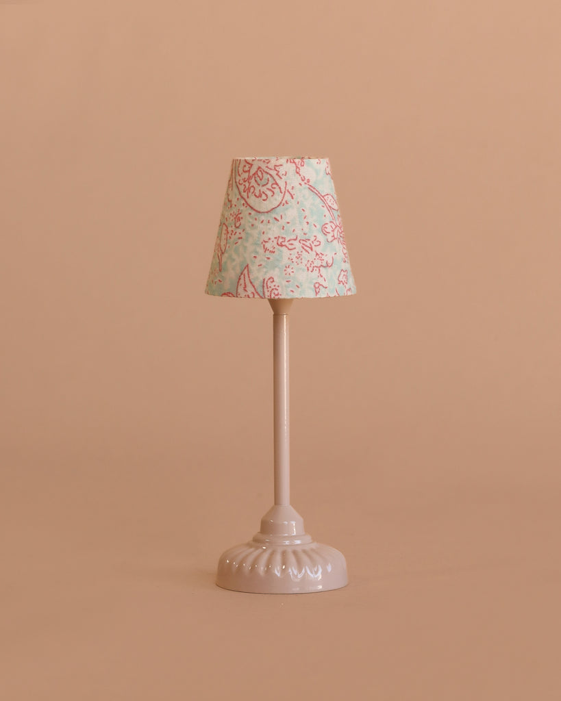A small table lamp with a pale pink base and floral-patterned shade sits beside a charming Maileg Nursery Set rocking horse, all arranged against a plain light brown background.