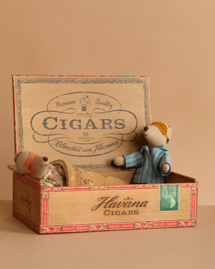 Two small mice from the Maileg Mouse Family Collection, one lying under a blanket and one sitting up in pajamas, are inside an open Maileg cigar box with "CIGARS" and "Havana" labels.