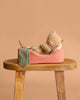 The Maileg Small Sleeping Kitten in Cozy Bed lies on its back in a pink pet bed with a green knit blanket, set on a wooden stool against a beige background.