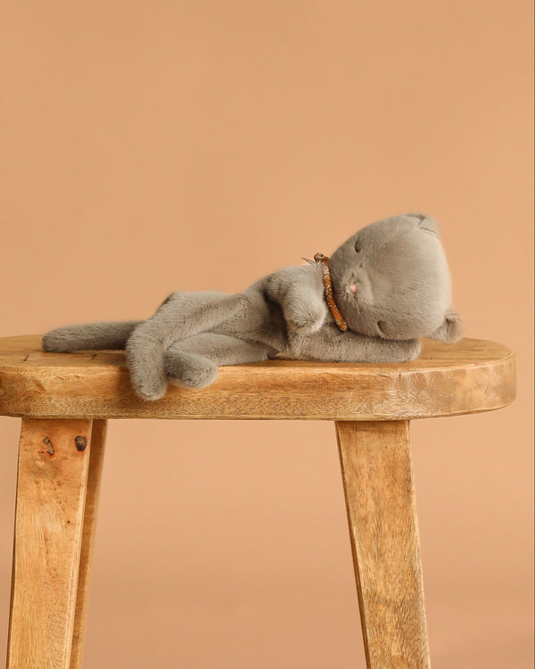 The Maileg Sleeping Kitten Plush, Small - Grey, rests on its side atop a wooden stool against a plain beige background.