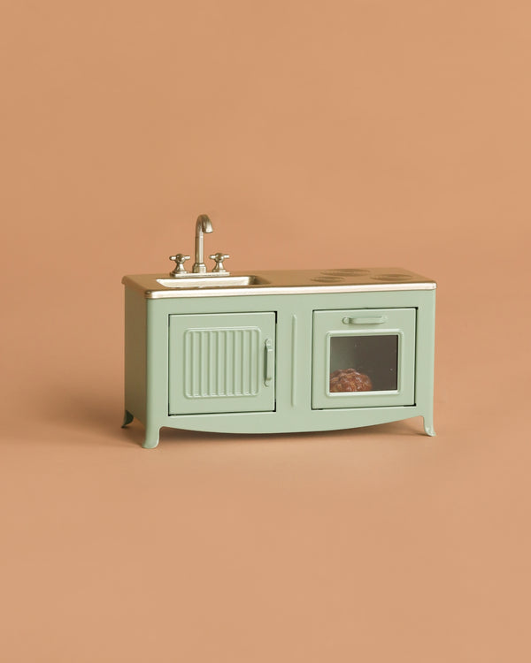 The Maileg Kitchen, Mouse - Mint is a small, pale green metal kitchen set with sink, faucet, and two cabinet doors (one with a window showing a tiny loaf of bread), perfect for any mouse family. Displayed on a plain beige background.