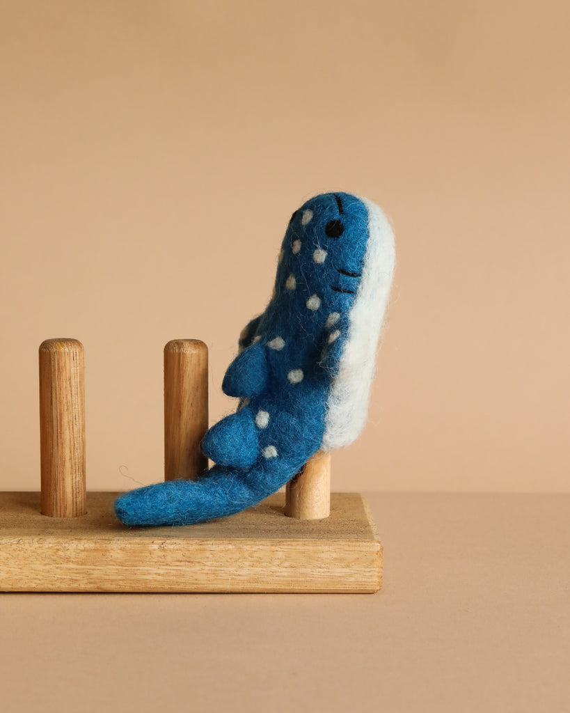 The Finger Puppet: Whale Shark, a blue and white felt plush with white spots, sits upright on a wooden rack with two vertical pegs against a plain beige background.