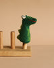 The Felt Gator Finger Puppet, featuring big eyes and a green felt design, is displayed on a wooden stand with three pegs against a beige background—ideal for imaginative play and storytelling.