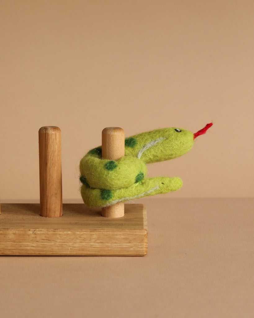 The Snake Finger Puppet, featuring green fabric with dark spots and a red tongue, wraps around a wooden peg on a small wooden base, inspiring creative storytelling and imaginative play.