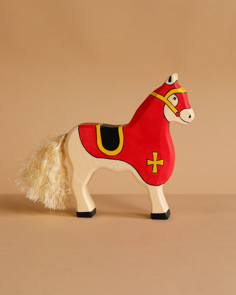The Holztiger Tournament Horse, Red ships in one week and features a handmade cream wood body, red saddle with yellow cross, black hooves, and cream yarn tail. Made from maple and beech wood. Shown against a beige background.