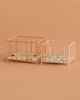 Two empty rectangular baby playpens with thin, vertical bars and patterned mattresses are placed side by side on a plain beige background. The Maileg Playpen, Micro – Dark powder is smaller than the other playpen beside it.