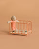 A small stuffed doll in a pink outfit stands inside the Maileg Playpen, Micro - Dark powder, featuring a patterned mattress and set against a plain peach background.