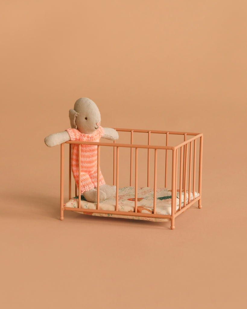 A small stuffed doll in a pink outfit stands inside the Maileg Playpen, Micro - Dark powder, featuring a patterned mattress and set against a plain peach background.
