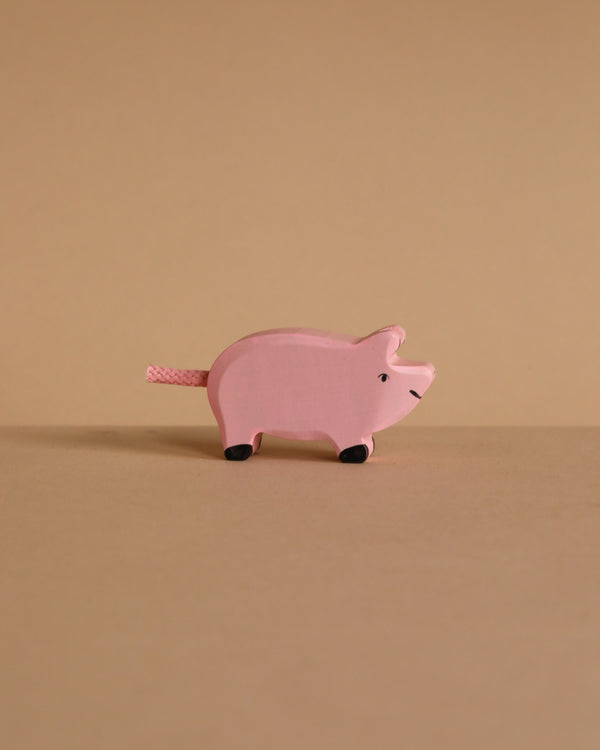 The Holztiger Piglet is a small, pink, handcrafted wooden toy with black feet, displayed on a beige surface against a matching background and exemplifying Holztiger’s renowned quality.