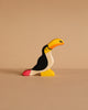 The Holztiger Toucan, a small handcrafted wooden figurine painted in black, white, yellow, orange, and pink, stands on a beige surface with a matching background. Ships in one week.