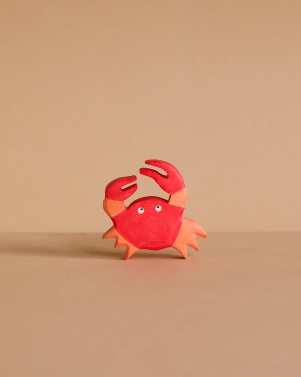 A small, red and orange cartoon crab with two raised claws and wide eyes stands on a light brown surface—just like the handcrafted Holztiger Crab, made in Europe and ships in one week.