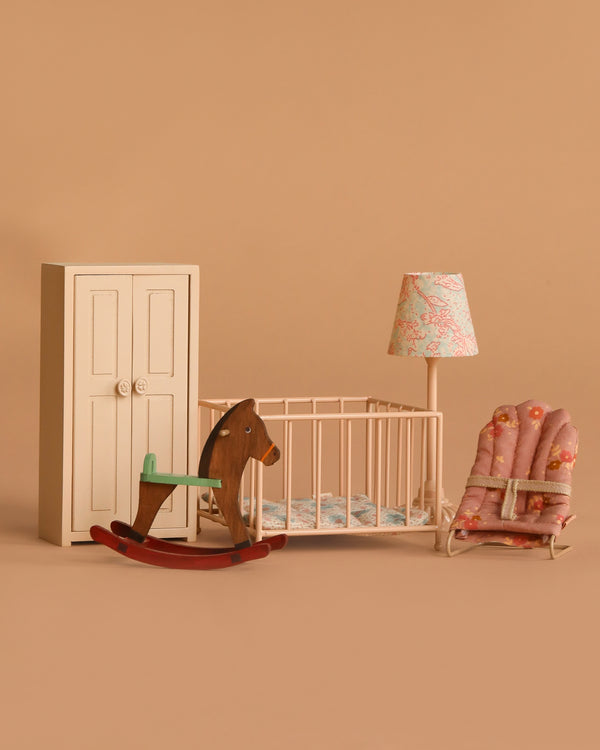 The Maileg Nursery Set includes a white wardrobe, wooden rocking horse mouse, crib with bedding, floral lamp, and pink floral-patterned baby rocker—adorable accessories for your baby mouse.