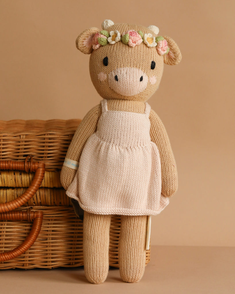 Cuddle + Kind Amelia The Cow is a hand-knit beige cow doll in a pale pink dress with a flower crown, posed before a wicker basket. This sustainable, fair trade toy makes a charming addition to any nursery.