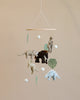 A nursery mobile featuring plush animals such as elephants and dinosaurs, accompanied by felt leaves and white orbs, hanging against a neutral backdrop.