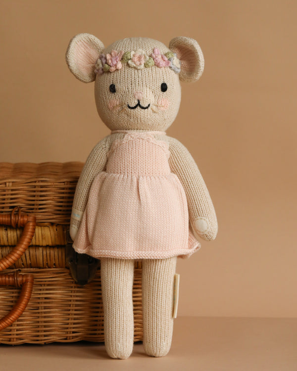 The Cuddle + Kind Emma The Mouse is a cream-colored, handmade knitted doll made from hypoallergenic materials, dressed in a light pink dress with a flower crown, shown standing before a wicker basket on a beige background.