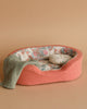 The Maileg Sleeping Bunny With Bed features a peach-colored pet bed with a floral interior. The bunny is nestled on a white pillow and green knitted blanket, crafted from recycled polyester, creating coziness against the warm beige background.