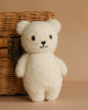 The Cuddle + Kind Baby Bouclé Bear - Ivory, a soft white plush toy with black eyes and a small smile, stands in front of a woven wicker basket on a beige background.