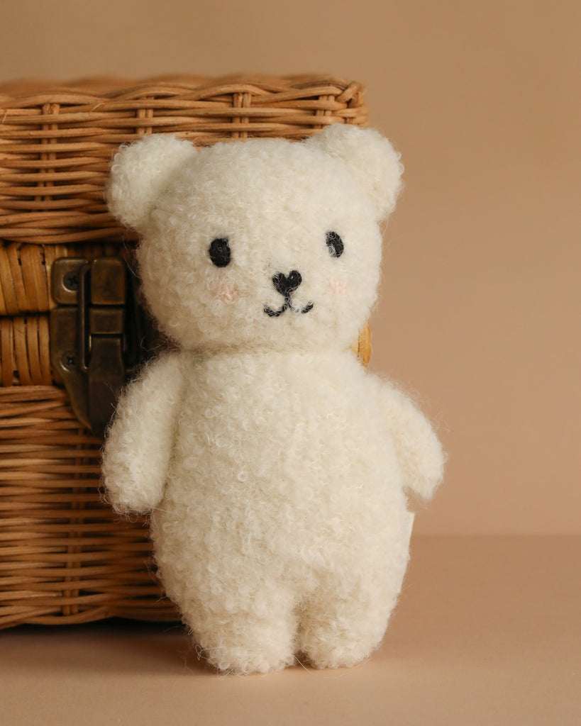 The Cuddle + Kind Baby Bouclé Bear - Ivory, a soft white plush toy with black eyes and a small smile, stands in front of a woven wicker basket on a beige background.