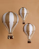 Three decorative handmade Hot Air Balloon - Gold ornaments, featuring alternating white and beige panels with small brown baskets, are suspended against a solid beige background—perfect for charming children’s room decoration.