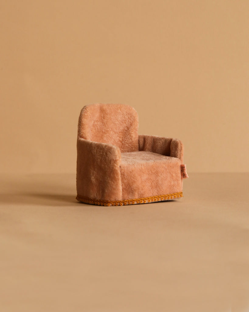 A soft pink armchair inspired by the Maileg Big Sister and Me Set sits on a matching beige surface with a beige background. The plush chair features low arms, a cushioned seat, and gold trim at the base.