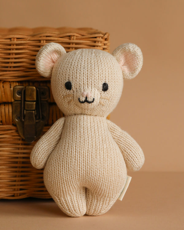 A beige, hand-knit Cuddle + Kind Baby Mouse with black eyes and a stitched smile sits upright by a woven wicker basket, crafted from natural Peruvian cotton yarn on a light brown surface.