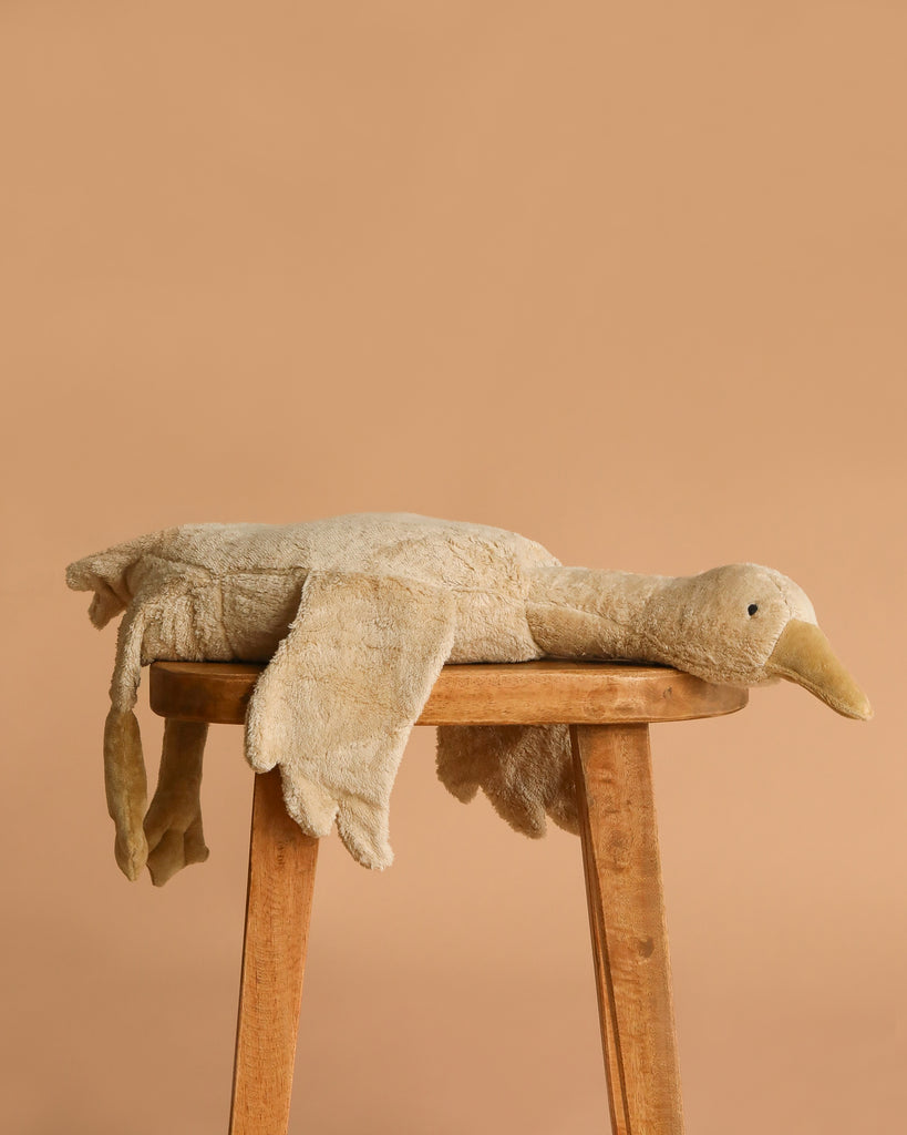 The Senger Naturwelt Cuddly Animal - Linen Goose, made with soft GOTS-certified cotton, rests on a wooden stool against a plain light brown background.
