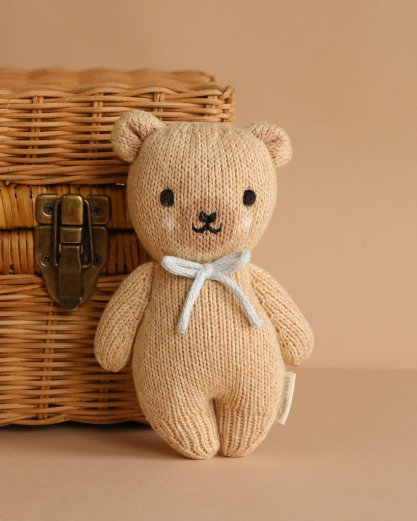 A small, knitted Cuddle + Kind Baby Honey Bear in Robin’s Egg Blue with a white ribbon around its neck sits in front of a wicker basket on a light brown surface and background.
