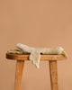 A worn, beige stuffed animal resembling the Senger Naturwelt Cuddly Animal - Linen Goose lies flat on its stomach on a wooden stool against a plain, light brown background.