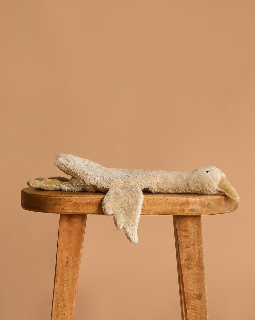 A worn, beige stuffed animal resembling the Senger Naturwelt Cuddly Animal - Linen Goose lies flat on its stomach on a wooden stool against a plain, light brown background.