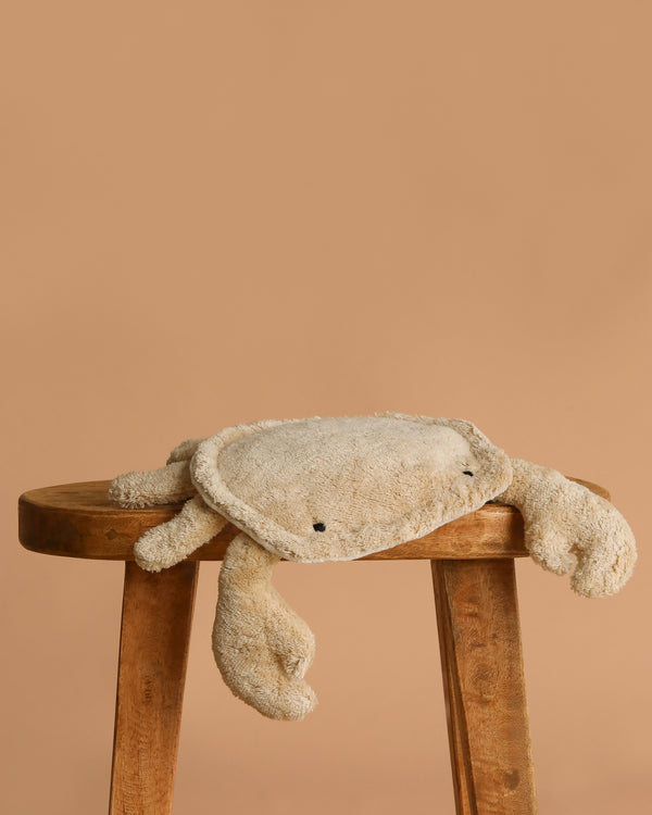 The Senger Naturwelt Cuddly Animal - Linen Crab, Small sits on a wooden stool against a light brown background—this soft plush toy is ideal for comfort and doubles as an organic cotton heat pad.